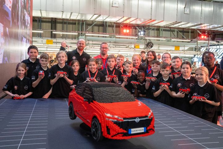 Hinter einem großen LEGO Modell eines Nissan Qashqai steht lächelnd eine Gruppe von Schulkindern mit kleinen Lego Qashqai in ihren Händen