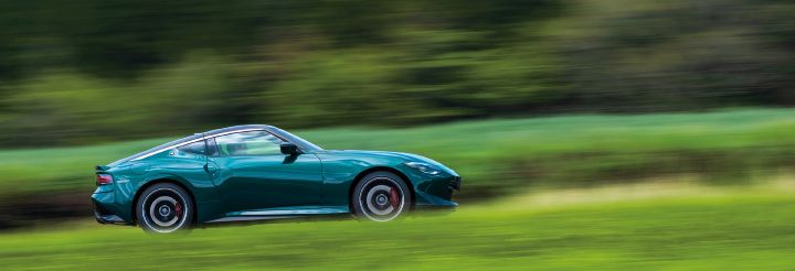 Unryu Green Fairlady Z captured in motion from the left-side profile using a panning shot. The vehicle faces toward the right side of the image, highlighting its long-hood, short-deck sports car proportions and muscular rear fenders. Black alloy wheels and red brake calipers are visible, while the surrounding grass and trees are motion-blurred to emphasize speed and movement. Set against a vibrant green natural landscape, the rich Unryu Green body color stands out, creating a dynamic image that showcases the Fairlady Z in motion.