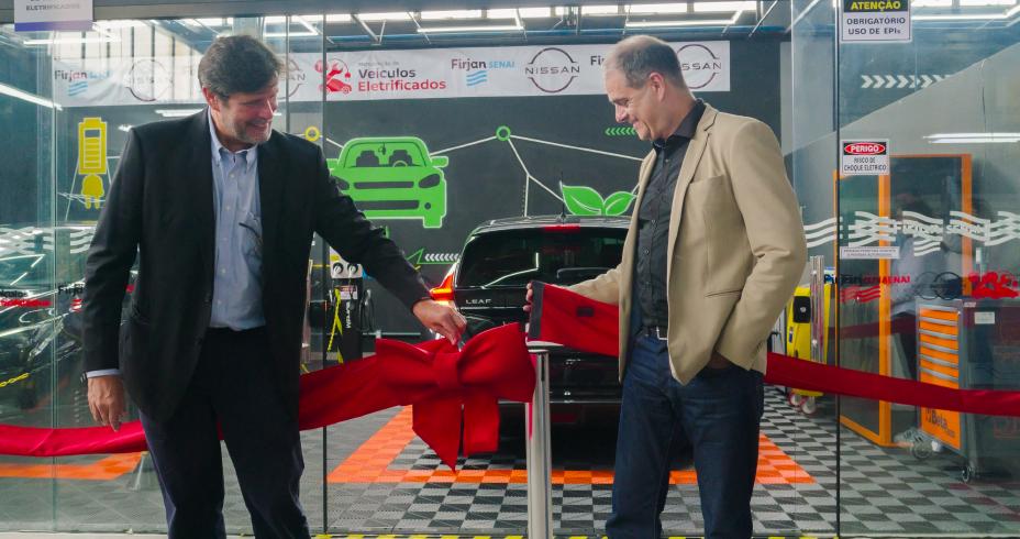 Imagem de dois homens brancos de terno um ao lado do outro cortando uma fita inaugural vermelha em frente a um veículo elétrico da Nissan preto