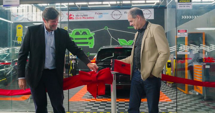 Imagem de dois homens brancos de terno um ao lado do outro cortando uma fita inaugural vermelha em frente a um veículo elétrico da Nissan preto