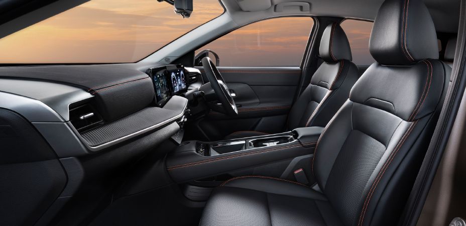 Interior view of the kicks rock creek photographed from the passenger side, focusing on the front seating area. The black-themed cabin features a steering wheel and digital meter display toward the front right, with a center display and center console located in the middle. The driver and front passenger seats are upholstered in black material, accented with lava red stitching on the headrests, seat bolsters, and center armrest. Matching stitching on the dashboard and door trims enhances both the premium feel and outdoor-inspired design.