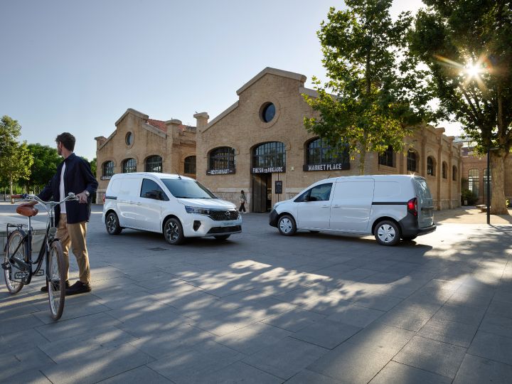 Vue latérale de deux Nissan Townstar EV Fourgon L2 blancs en face à face avec au premier plan un homme faisant dos à la caméra et tenant son vélo, un marché fermé est également présent en arrière plan.