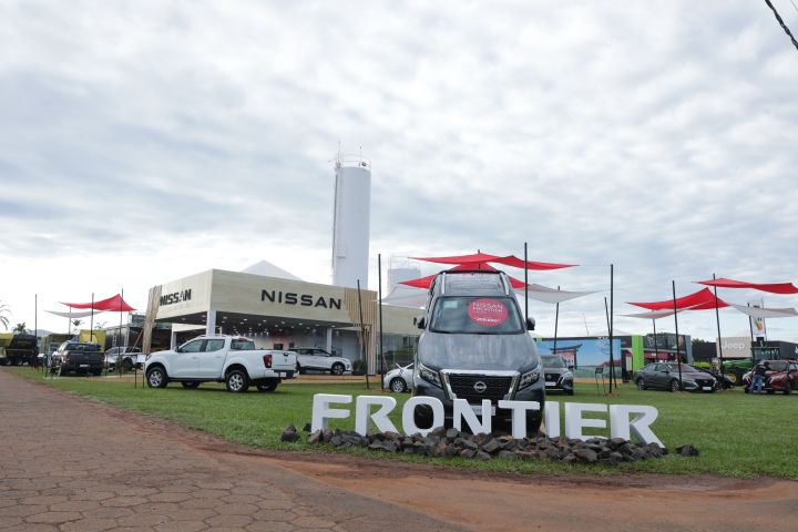 Estande da Nissan na Tecnoshow Comigo 2025 em Rio Verde (GO), com destaque para a picape Nissan Frontier posicionada em frente ao letreiro "FRONTIER" sobre pedras decorativas. Ao fundo, o estande moderno exibe outros veículos da marca, sob estruturas com coberturas vermelhas e brancas. Um cartaz no para-brisa anuncia condições especiais de venda da versão Pro-4X