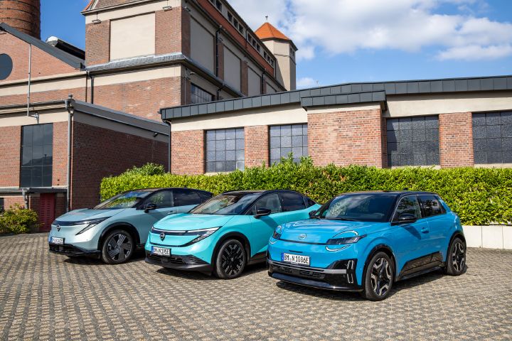 Vor einem Backsteingebäude und einer grünen Hecke stehen nebeneinander die drei Nissan EV-Modelle Nissan Ariya, Nissan Leaf und Nissan Micra, seitlicher Blick auf die Gruppe mit dem Micra im Vordergrund