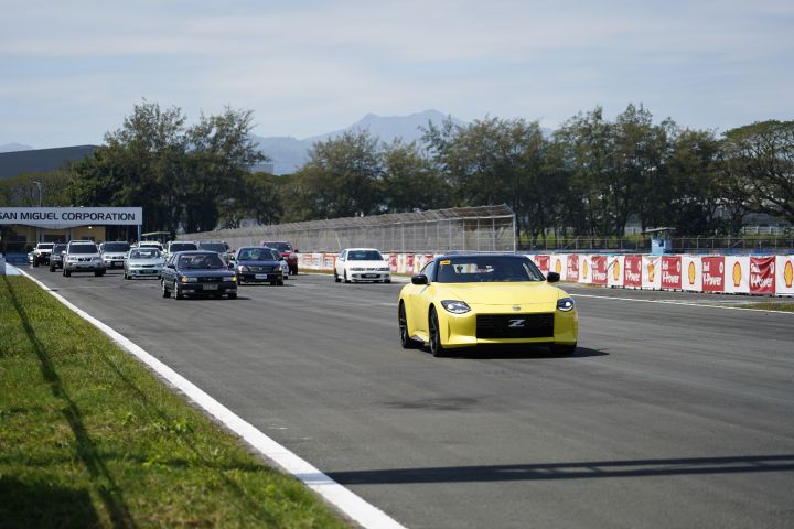 A yellow Nissan Z sports car leads a lineup of various vehicles driving on a race track, with trees, mountains.