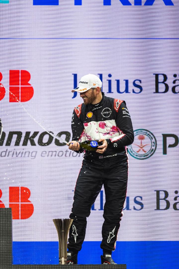 Piloto da Equipe Nissan de Fórmula E sorrindo no pódio, segurando uma garrafa de champanhe aberta