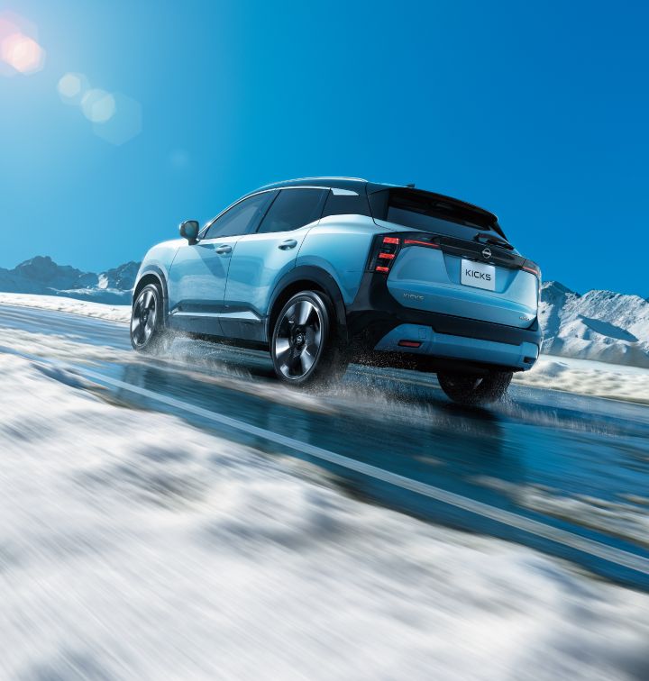 A bluish Nissan Kicks driving on a snowy road. The vehicle moves to the right, captured from a rear perspective, with a snowy landscape and blue sky in the background.
