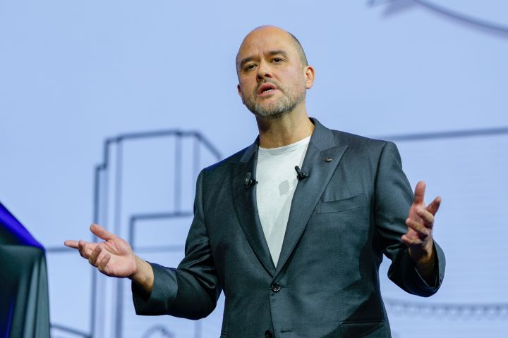 On stage, CEO Ivan Espinosa stands in a suit, gesturing with both hands while delivering a presentation. The background features a white screen with simple line art.