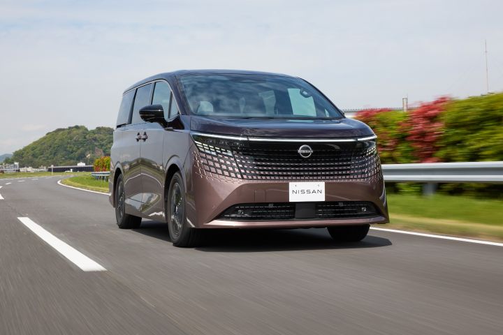 新型エルグランドが、左から右へ向かってカーブした道路を走行している。車両は右前方へ進む斜め前からの低いアングルで捉えられ、大型のブラックグリルと横一文字に伸びるLEDライトが強調されている。背景には淡い青空と薄い雲、緑の丘やガードレール、赤い花が植えられた植栽帯が広がり、周囲はモーションブラーで表現されて走行中のスピード感が際立っている。