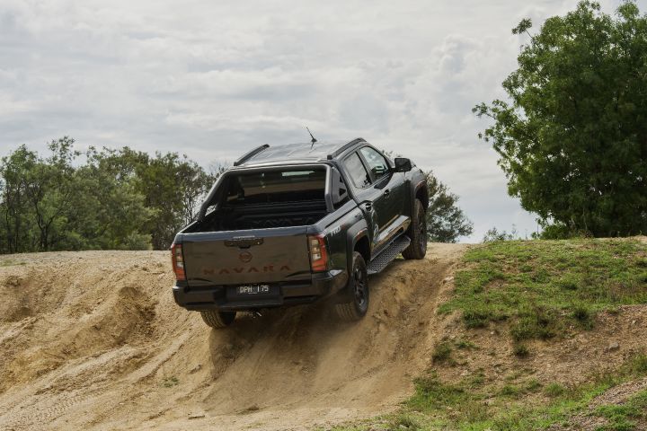 2026 Nissan Navara PRO-4X finished in Boulder Grey shown rear three‑quarter climbing with light dust, open tub beneath the sports bar and right‑rear wheel compressed.