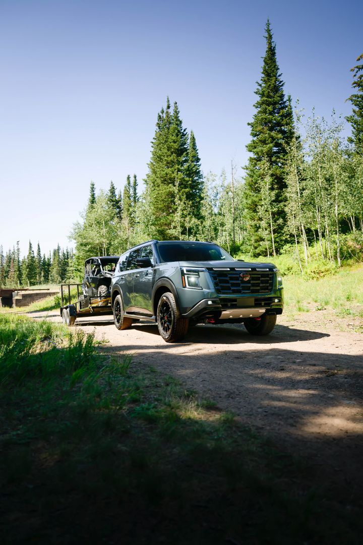 Front right 3/4 view of a green 2025 Nissan Armada on a dirt road towing a trailer, pines and brush are in the background.