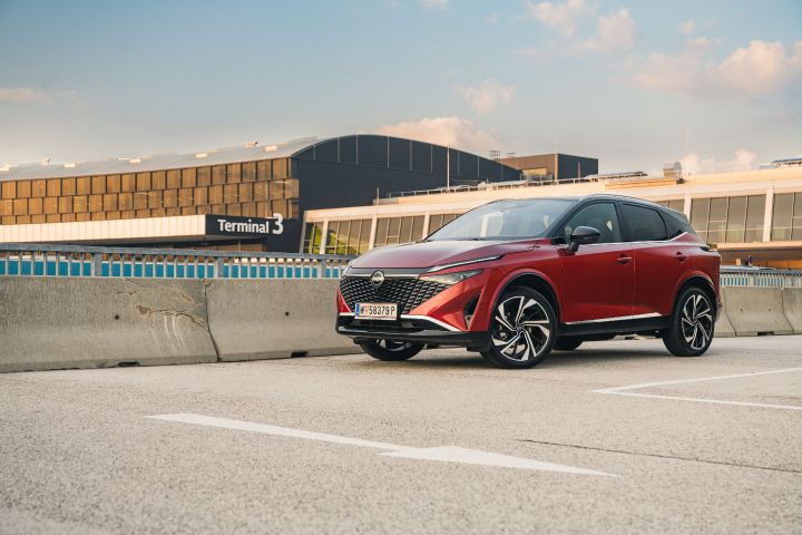 Ein Nissan Qashqai in Fuji Sunset Red steht auf einem Parkhausdach vor einem Flughafenterminal