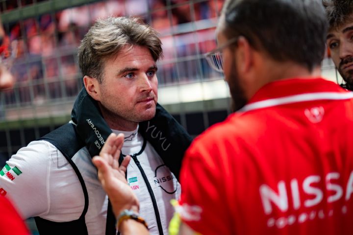 Oliver Rowland, der einen weißen Rennanzug trägt, steht mit ernstem Blick in der Startaufstellung und unterhält sich mit mehreren Nissan Teamkollegen, die rote Teamkleidung tragen.