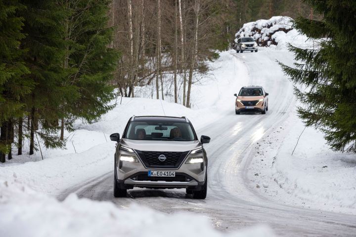 Silberner Nissan X‑Trail e‑Power e‑4ORCE fährt auf schneebedeckter Straße vor dem Elektroauto Nissan Ariya e‑4ORCE in Akatsuki Copper mit Dach und Außenspiegel in Pearl Black
