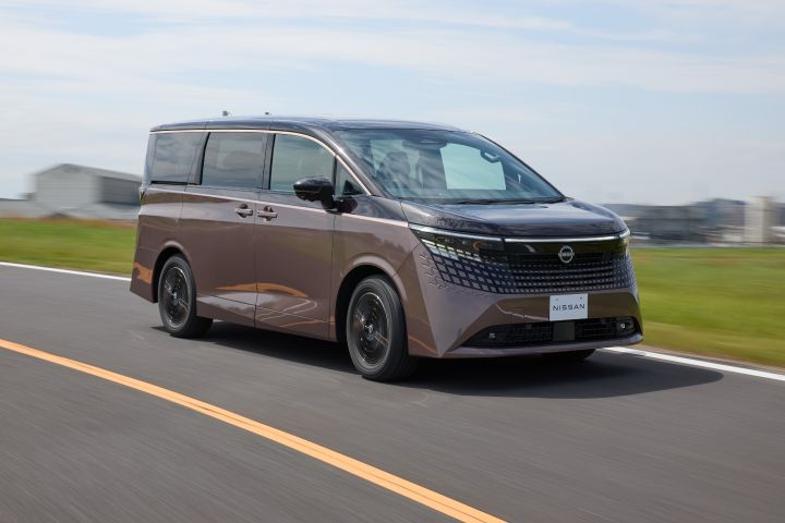 新型エルグランドが、左から右へ向かって直線的な舗装道路を走行している。車両は右前方へ進む斜め前からのアングルで捉えられ、ブラックの大型グリルとシャープなLEDヘッドライトが際立つ。空は淡い青色で薄い雲が広がり、道路脇には緑の芝生と低い建物が見え、背景は流れるようなブラーでスピード感が表現されている。