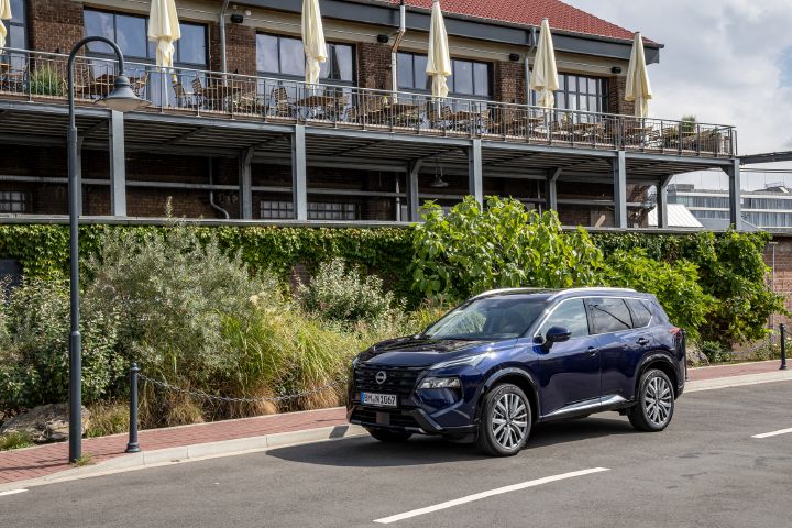 Vor einem Gebäude mit Backsteinfront und mehreren Balkonen steht ein dunkelblauer Nissan X‑Trail