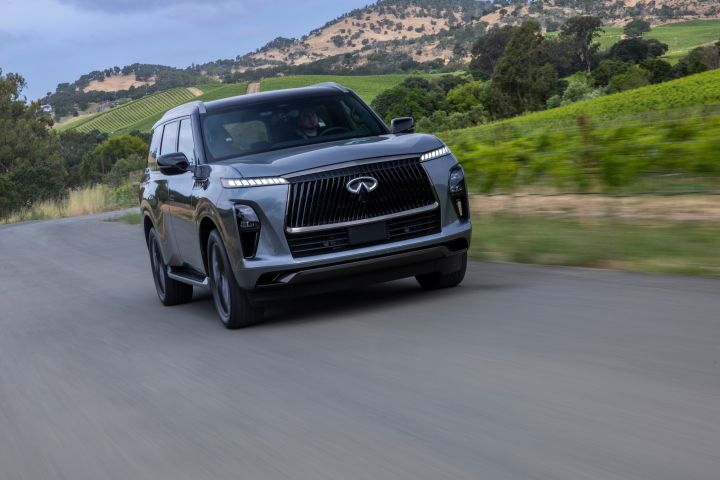 なだらかな丘とブドウ畑を背景に田舎道を走るダークグレーの INFINITI QX80 SUV