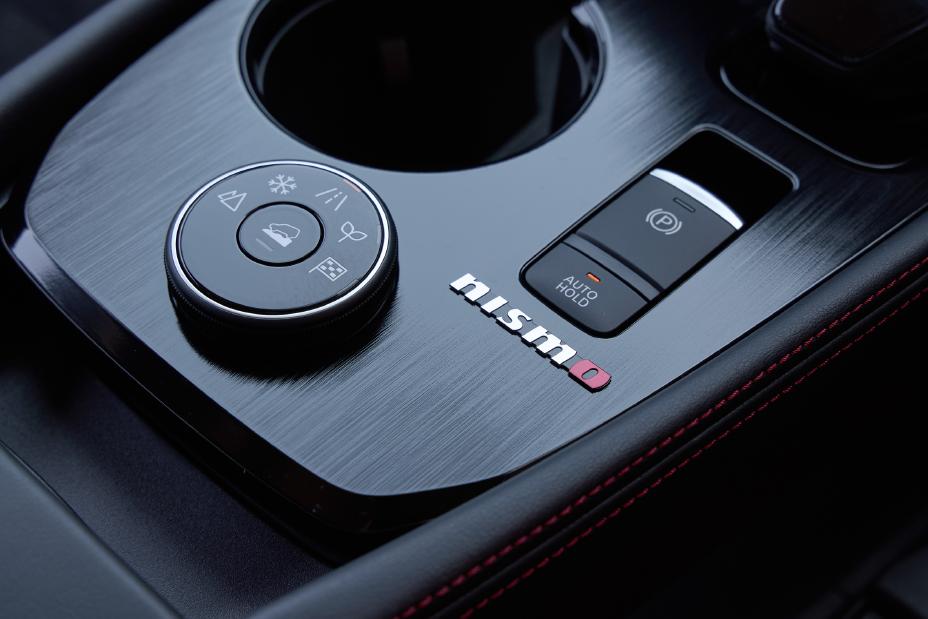 Center console of a Nissan X-Trail NISMO. The area around the gear shift features a circular control knob with various settings, an electronic parking brake button, and the “NISMO” logo displayed in white and red text.