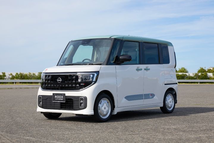 The new Nissan Roox kei car is parked in a lot under a clear blue sky. Its white body reflects natural daylight, with paved ground and other vehicles visible in the surroundings.