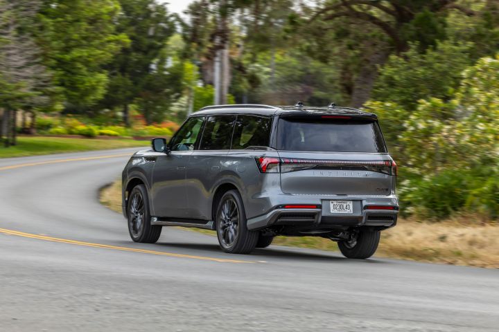 緑を背景に走行するダークグレーの INFINITI QX80 SUV の後方 3/4 ビュー