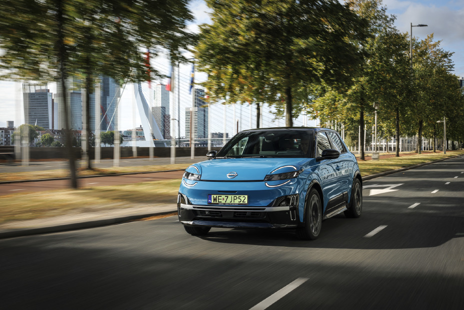 Niebieski elektryczny Nissan Micra jedzie miejską ulicą w dynamicznym ujęciu, z panoramą miasta w tle.