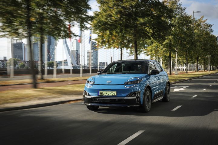 Niebieski elektryczny Nissan Micra jedzie miejską ulicą w dynamicznym ujęciu, z panoramą miasta w tle.