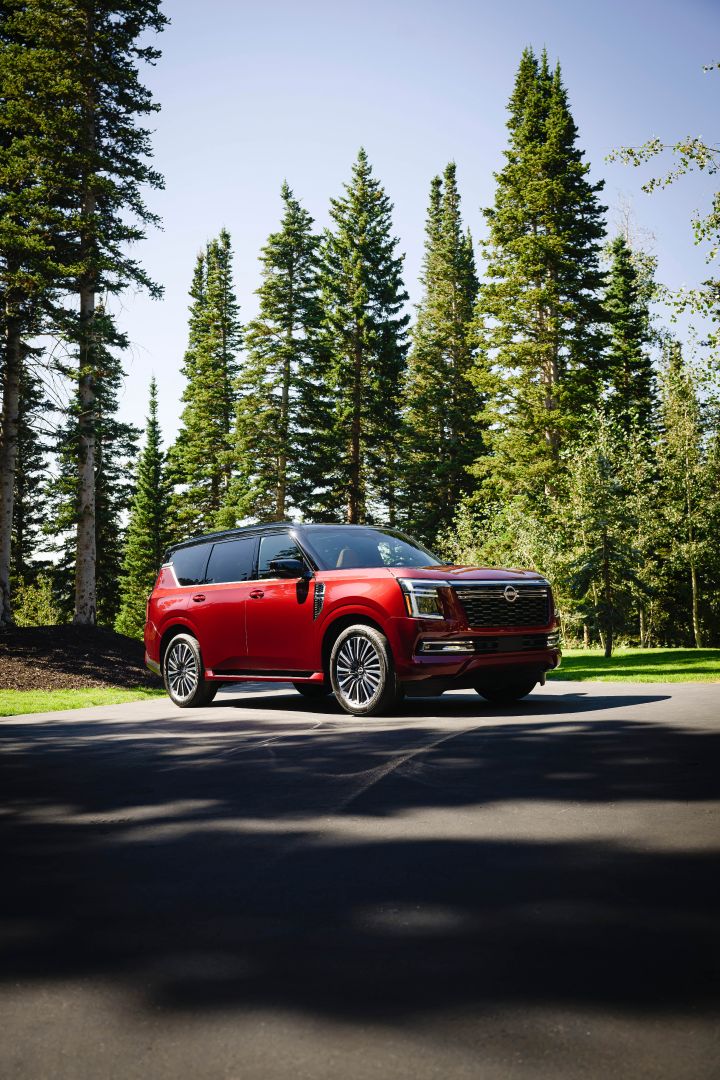 Front right 3/4 view of a red 2025 Nissan Armada parked in front of tall pine trees and blue skies.