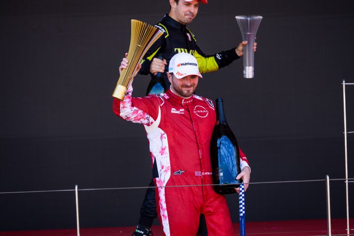 Nissan Formel E Fahrer Oliver Rowland hält in der rechten Hand den Siegerpokal des E-Prix von Monaco hoch und in der linken eine Flasche Champagner.
