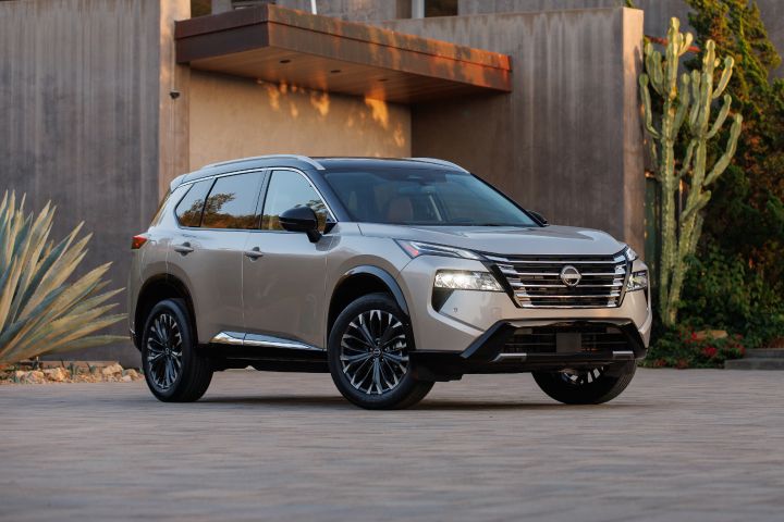 Front right ¾ view of a silver 2024 Nissan Rogue parked in front of a cement building with arid landscaping alongside.