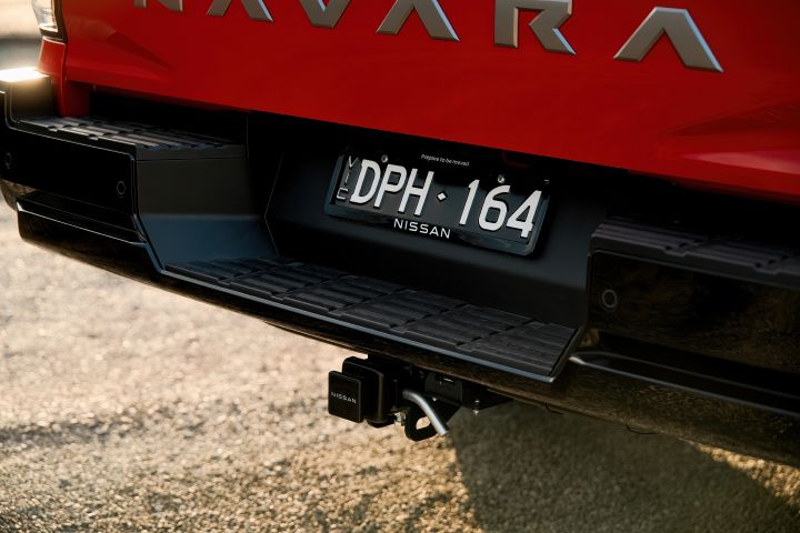 Close-up of the rear bumper and tow bar on a 2026 Nissan Navara ST‑X in Outback Red, showing the trailer hitch, wiring socket, step surface, rear number plate, and lower tailgate edge.