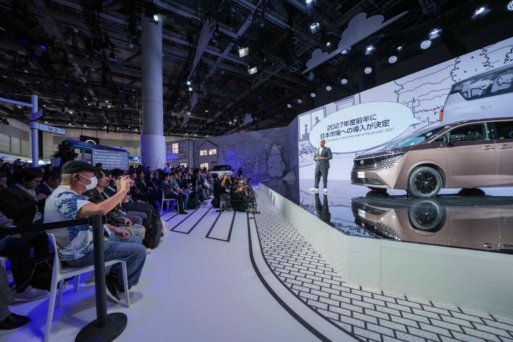 On the stage, the all-new Elgrand in Fuji Dawn and Shigoku two-tone colors is positioned diagonally, facing slightly to the left. Next to the vehicle stands CEO Ivan Espinosa in a suit, delivering a presentation. The background features a white screen. In the foreground, members of the press are seated, taking photos with cameras.