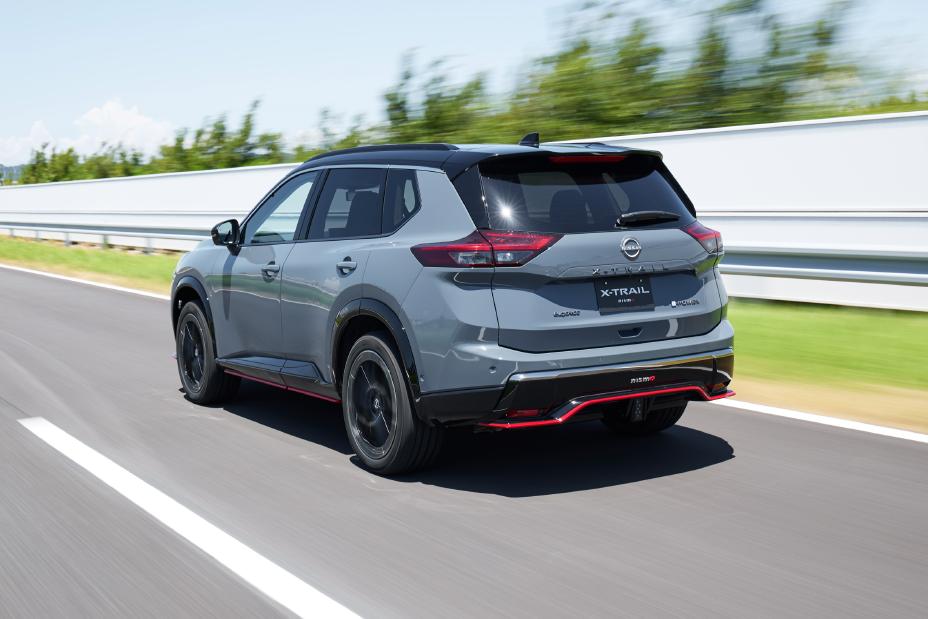 A grey Nissan X-Trail NISMO with black and red accents is driving on a multi-lane road. There is a barrier on the right side, and the background features trees and greenery.