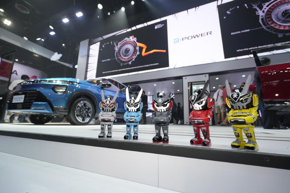 A row of five small, colorful robot figurines styled like mascots is displayed on a platform at an auto show booth. Behind the figures, a blue Nissan vehicle is partially visible, along with attendees walking around the exhibit. Large digital screens in the background showcase Nissan’s e‑POWER technology, including a graphic of an electric motor and drivetrain diagram. Bright overhead lights illuminate the modern exhibition space.