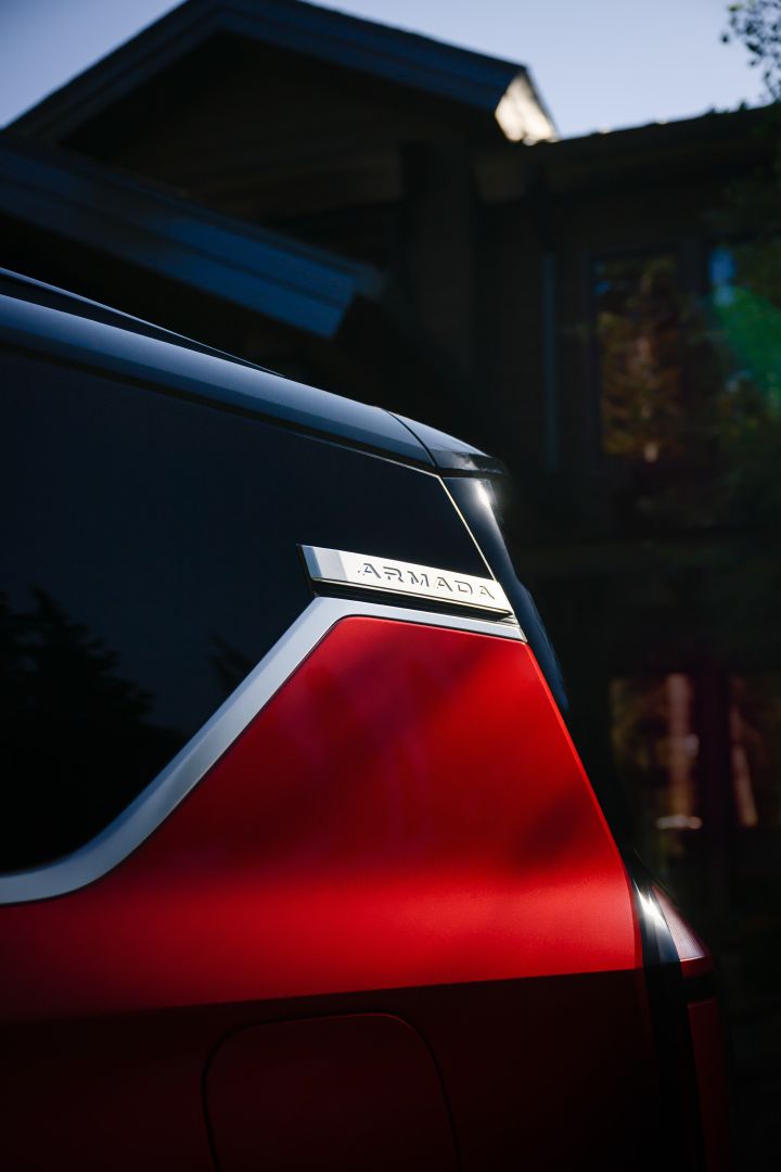 Close up of the top half of a red 2025 Nissan Armada, the Armada logo can be seen in the upper right and a building behind.