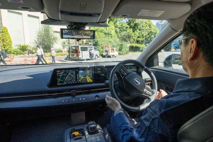 Innenansicht eines Nissan Ariya. Der Innenraum ist in Schwarz gehalten und verfügt über ein großes Armaturenbrett-Display, auf dem ein Navigationssystem angezeigt wird. Eine Person sitzt auf dem Fahrersitz, ohne das Lenkrad festzuhalten und blickt nach vorne. Im Hintergrund sind Gebäude und Bäume zu sehen, die einen Blick auf die Außenumgebung ermöglichen.