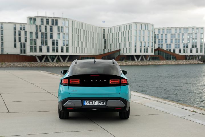 Frontale Heckansicht eines türkisen Nissan LEAF mit schwarzer Heckklappe, im Hintergrund ist die Front eines modernen weißen Gebäudes zu sehen