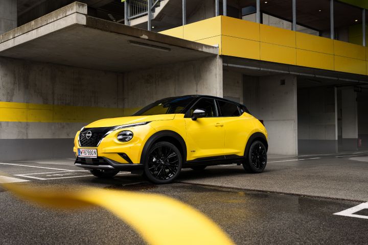Gelber Nissan Juke steht schräg von vorne vor einem grauen Gebäude mit mit gelben Elementen, im Vordergrund sind verschwommen gelbe Bänder zu sehen