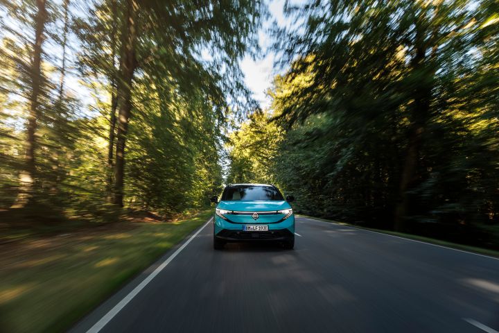 In der Bildmitte frontale Fahraufnahme eines türkisen Nissan LEAF, die Straße führt durch einen Wald