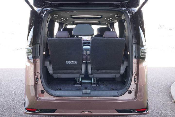 新型エルグランドのリアハッチを開けた状態で、車両後方から車内を正面に捉えたビュー。3列シートの後ろ側が見えており、ブラックのシート背面とヘッドレストが左右対称に配置されている。床面にはカーペットとシート固定用のラッチやストラップが見え、3列目シートの背面には折りたたみ方法を示すラベルが貼られている。前方には2列目シート越しにダッシュボードと中央ディスプレイがわずかに見える。周囲は明るい屋外環境で、外光がラゲッジスペースに差し込み、広く実用的な荷室空間が強調されている。