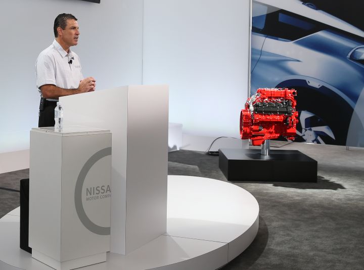 a man giving a speech in a podium next to a red vehicle engine