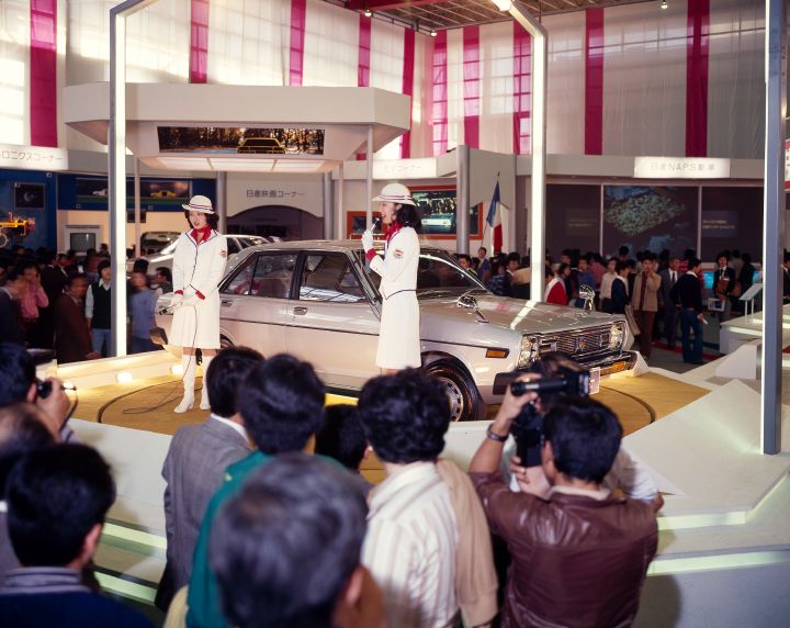 a crowd of onlookers viewing a presentation of a vintage beige nissan vehicle at an auto show
