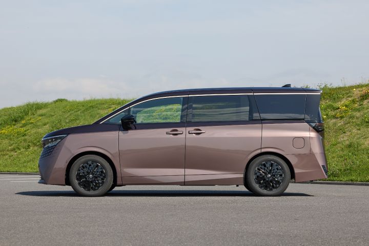 新型エルグランドが、左から右へ向かう側面ビューで静止している。車両は右向きで、ブラックルーフとのツートーンカラー、直線的で伸びやかなボディライン、ブラックの多スポークホイールが強調されている。背景には緑の草地と小さな黄色い花が広がる土手があり、上空には淡い青空が広がる明るい日中の屋外環境で、前景には灰色の舗装路面が広がっている。