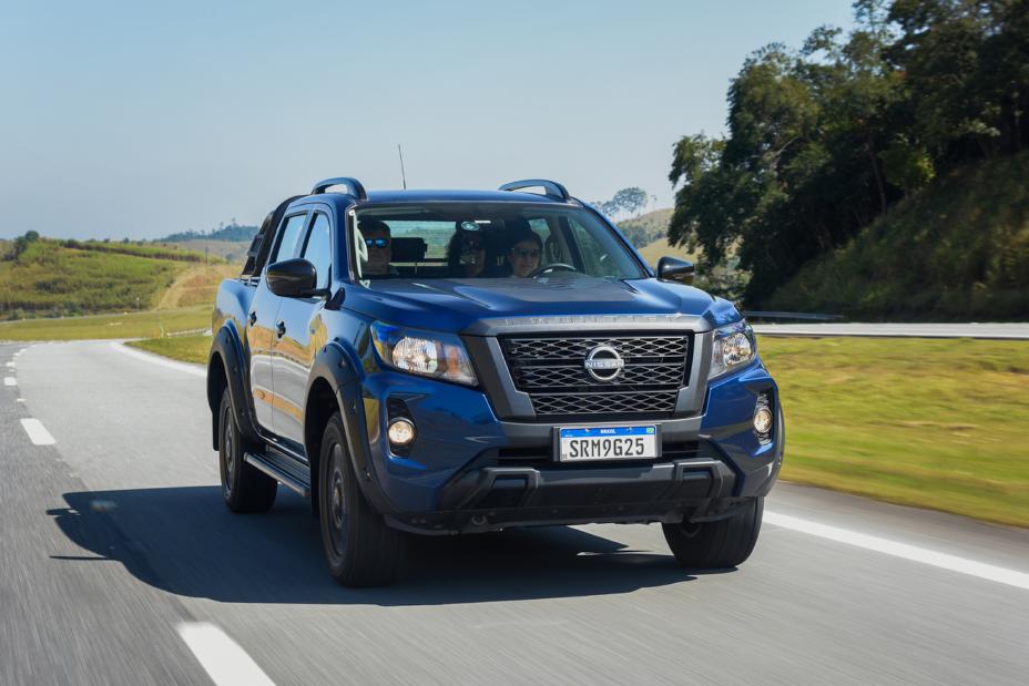 Uma picape Nissan Frontier Attack 2025 na cor azul trafega por uma estrada asfaltada durante o dia. O veículo possui uma grade frontal preta, faróis modernos e detalhes escurecidos. No interior, três ocupantes são visíveis através do para-brisa, enquanto a paisagem verde e montanhosa compõe o fundo da imagem.