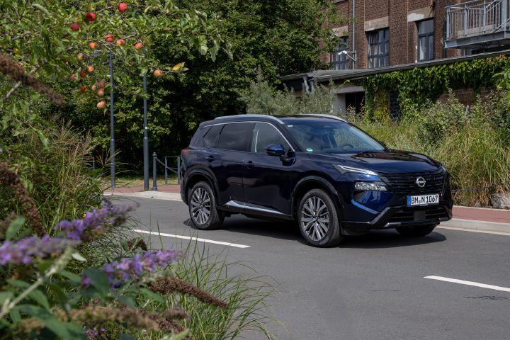 Blick von schräg vorne auf einen parkenden dunkelblauen Nissan X‑Trail, in den linken Bildvordergrund ragen grüne Pflanzen