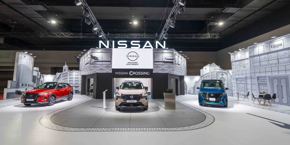 Wide view of a Nissan exhibition booth featuring three vehicles—red, white, and blue—displayed on circular platforms under a large “NISSAN” sign, with a modern, brightly lit showroom design and surrounding display panels.