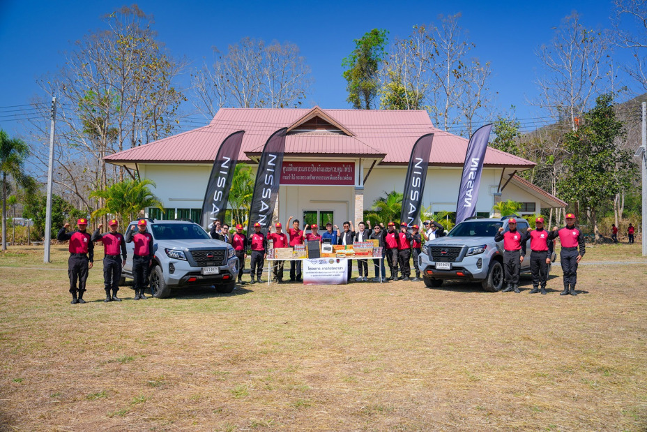 A group of people standing outdoors in front of a small building with a banner, posing between two vehicles during an organized event.