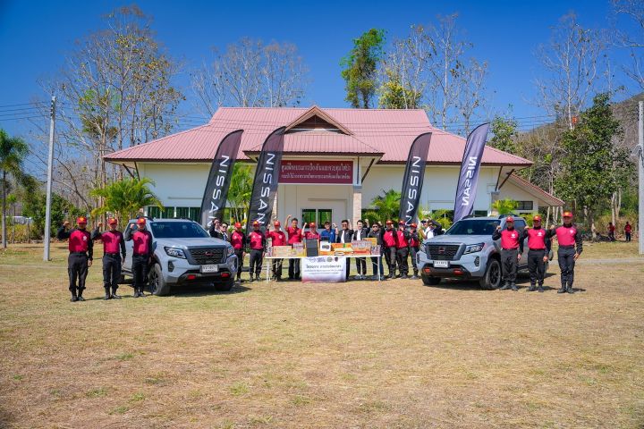 A group of people standing outdoors in front of a small building with a banner, posing between two vehicles during an organized event.