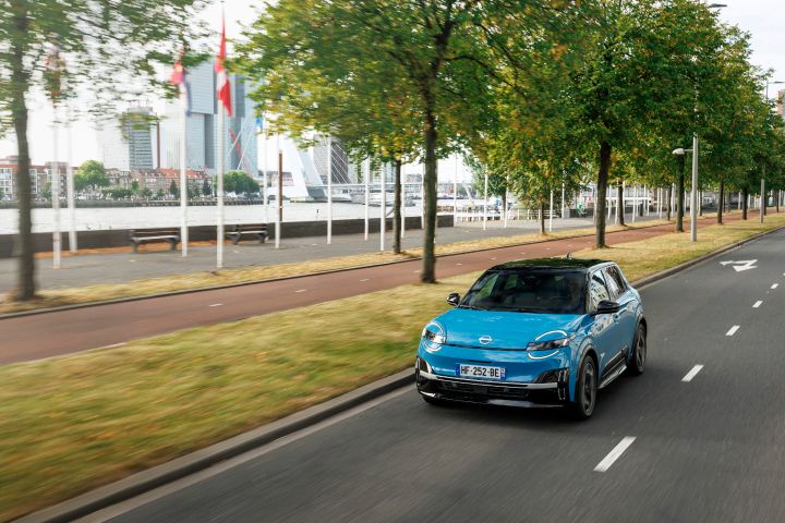 Blick von schräg oben auf die Front eines blauen Nissan Micra, der eine Straße in Rotterdam entlangfährt, im Hintergrund ein Radweg und Bäume