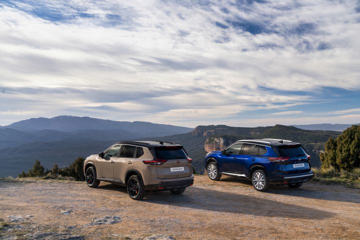 Zwei neue 2026 Nissan X‑Trail stehen auf einer Lichtung mit Blick auf eine bergige Landschaft unter einem teilweise bewölkten Himmel. Die Fahrzeuge, ein N-Trek in der Karosseriefarbe Baja Storm und ein Tekna in der Karosseriefarbe Universal Blue, stehen nebeneinander mit der Rückseite zum Betrachter und betonen so ihr Heckdesign und die raue Outdoor-Umgebung.
