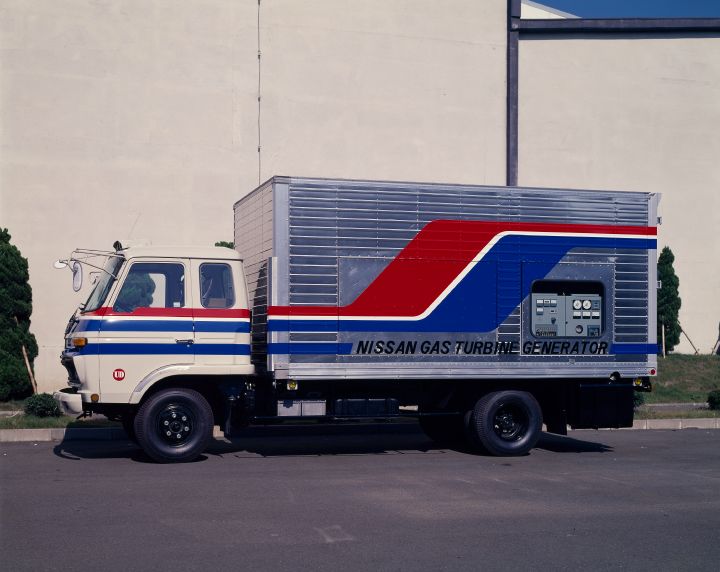 a movable gas turbine truck with a prominent red and blue stripe running across the side of the truck. The truck is parked outside of a large building and in large black letters has the text Nissan Gas Turbine Generator written across it.